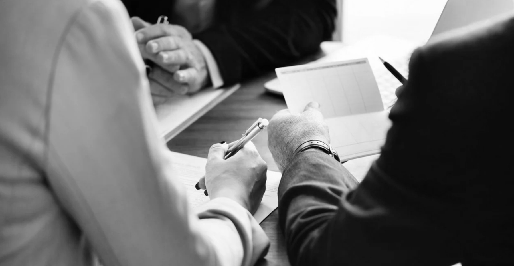 A black and white image depicting a group of individuals gathered around a table, engaged in conversation.